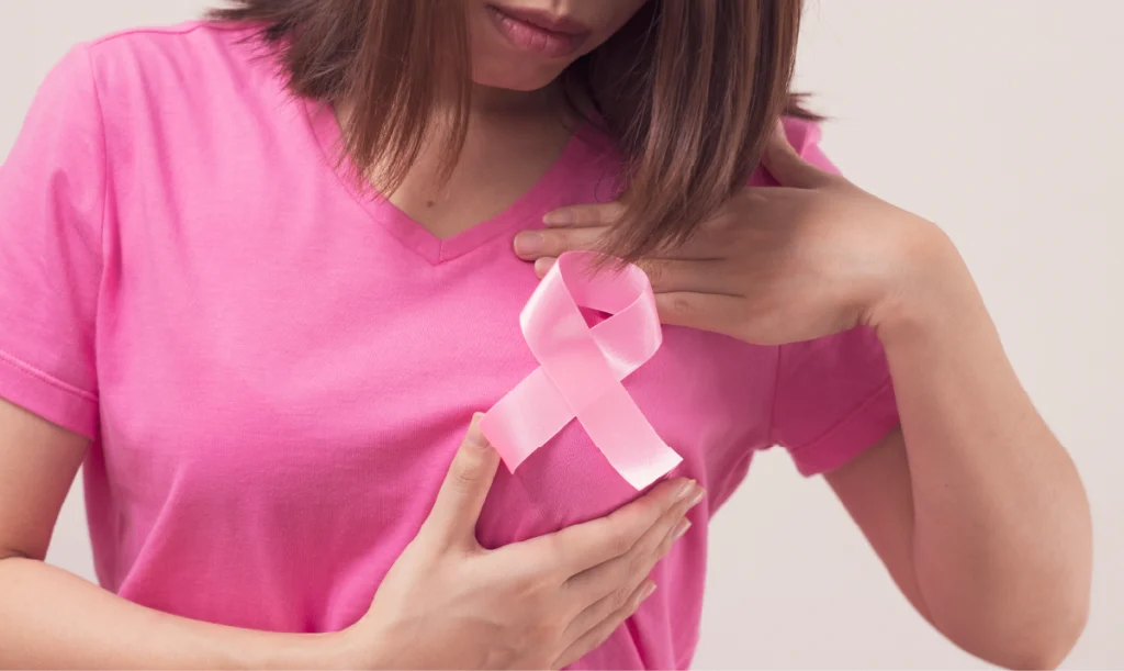 A woman in a pink shirt holds a pink ribbon over her chest, a global symbol of breast cancer awareness representing femininity, compassion, and hope.
