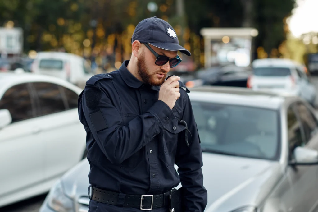 Law enforcement officer wearing a black uniform and speaking into a radio beside a vehicle.